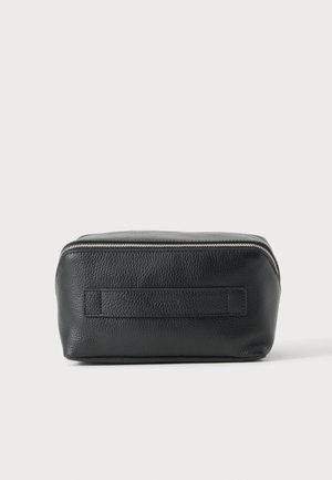 Trousse de toilette en cuir noir texturé avec poignée horizontale et fermeture éclair argentée sur le dessus, sur un fond clair uni.