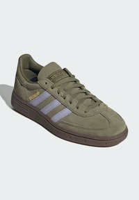 Sneakers de camurça verde-oliva com três riscas lilás, sola de borracha rígida castanha texturizada e marcação dourada na língua. Design clássico.