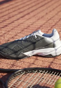Sort-hvid tennissko på rød lerbane ved siden af tennisracket og bold med netskygge, der overlapper scenen.