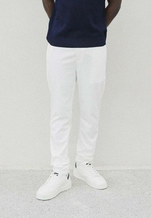 Pantalons en mélange coton blancs avec une coupe slim, présentant une texture douce et des poches latérales, associés à des baskets blanches et un pull marine.
