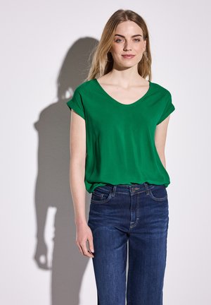 Grön, kortärmad, V-ringad blus i slätt tyg, ihop med mörkblå denimjeans med klassisk passform och synliga söm detaljer.
