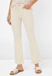 Beige flared jeans met een rafelige zoom, gemaakt van soepele stof. Gecombineerd met witte slidesandalen, met een open teen en banden.