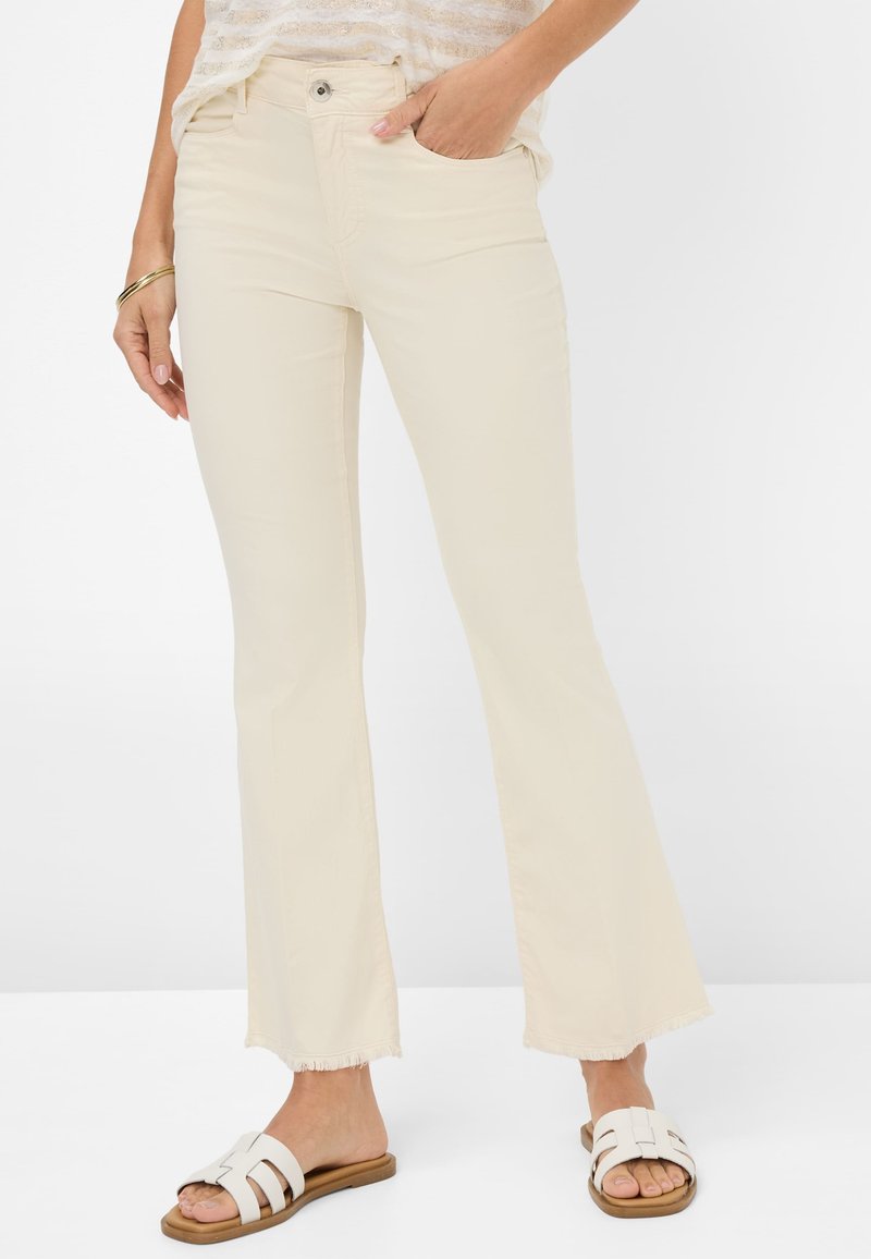Beige flared jeans met een rafelige zoom, gemaakt van soepele stof. Gecombineerd met witte slidesandalen, met een open teen en banden.