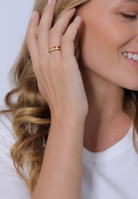 Goldring mit roten Akzenten an einer Hand, mit glatten, ineinander verschlungenen Bändern. Model hat welliges, blondes Haar und trägt ein weißes Oberteil.