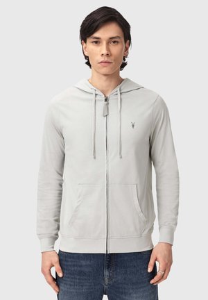 Mann trägt hellgrauen Kapuzenpullover mit Reißverschluss, Fronttaschen und Kapuze, kombiniert mit blauen Jeans, steht vor einfachem Hintergrund.