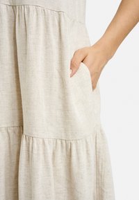 Helles beige Leinenkleid mit strukturiertem Finish, das ein schichtendes Design und eine Seiten-tasche aufweist. Der Stoff wirkt weich und atmungsaktiv.