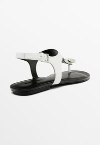 Hvid lædersandal med en sort sål, der har en spænde-strop og en rund detalje på oversiden. Glat tekstur og minimalistisk design.