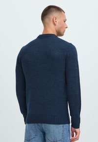 Mann med kort hår iført en marineblå genser og lyseblå jeans, stående og vendt bort fra en ensfarget lys bakgrunn.