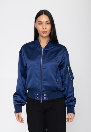 Vrouw met lang donker haar die een glanzend marineblauw bomberjack draagt met een rits aan de voorkant en een ritssluiting in de linker mouw, staand tegen een witte achtergrond.