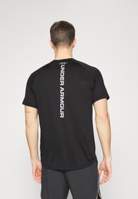 Svart sportt-shirt med kort ärm och rund hals, med en silvrig "Under Armour"-logotyp vertikalt längs ryggen. Mjuk, lätt material.