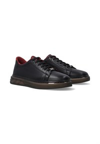Paar schwarze low-top Ledersneaker mit schwarzen Schnürsenkeln, rotem Innenfutter und durchsichtigen braunen Gummisohlen auf weißem Hintergrund.