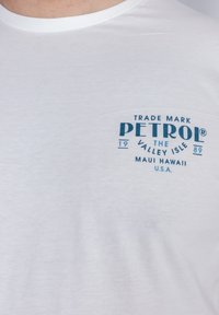Bílý bavlněný tričko s modrým grafickým potiskem na levé straně hrudi, na kterém je text "Petrol, The Valley Isle, Maui Hawaii, U.S.A."