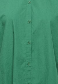 Green button-up shirt made of smooth fabric, featuring a collar, six buttons down the front, and a solid color without patterns or textures.