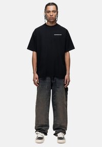 Zwarte t-shirt met korte mouwen en witte tekst op de borst, gecombineerd met losse donkerdenim jeans en witte sneakers.