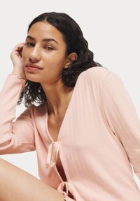 Roze geribde cardigan met een V-hals, voorzien van kleine strikjes in het midden en bij de manchetten. De stof heeft een zachte textuur en een subtiel verticaal patroon.
