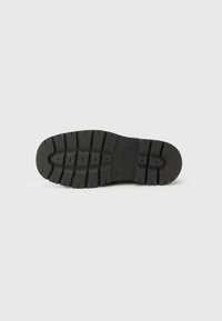 Semelle de chaussure en caoutchouc noir avec un motif de bande de roulement texturé, présentant des rainures profondes et une zone d'orteil arrondie. Comprend un logo rectangulaire en relief.