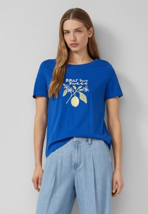 Frau mit langen hellbraunen Haaren, die ein blaues T-Shirt mit dem Schriftzug "sour but sweet" und Zitronengrafik trägt, kombiniert mit hellblauen Jeans.