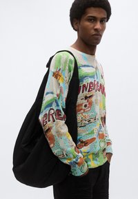 Πολύχρωμο πλεκτό πουλόβερ με ποικιλία σχεδίων και εικόνων, με το κείμενο «ΑΘΡΗΤΟΣ». Μοντέλο που φορά μαύρη τσάντα στον ώμο.