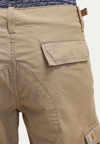 Pantaloni cargo beige in tessuto testurizzato, con taglio dritto, tasca laterale e dettagli di cucitura. Include un logo del marchio sulla tasca posteriore.