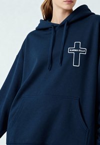 Felpa blu navy con tasca a marsupio frontale, caratterizzata da testo ricamato in bianco "KARMA KILLS" e design incrociato sulla parte sinistra del petto.