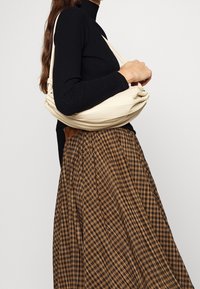 Beige slouchy tas met geplooide details, gedragen over een zwart geribbeld sweater, gecombineerd met een bruine geruite rok met plooien.