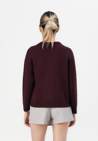 Žena s blond vlasy staženými do culíku, oblečená v tmavě fialovém svetr s pleteným vzorem a světlých šortkách, stojící na bílém pozadí.