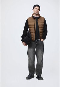 Barna steppelt mellény fekete hosszú ujjú póló felett, szürke farmernadrággal és fekete tornacipővel párosítva. Fekete táskával a kezében áll egy semleges háttér előtt.