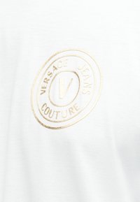 Λευκό ύφασμα με ανάγλυφο χρυσό οβάλ λογότυπο που γράφει "Versace Jeans Couture" περιβάλλοντας ένα κεντρικό γράμμα "V".