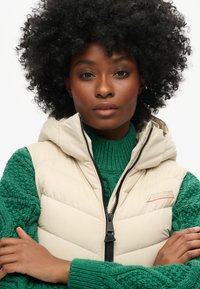 Doudoune beige avec capuche, fermeture éclair à l'avant et détails en broderie rouge, superposée sur un pull en tricot vert texturé.