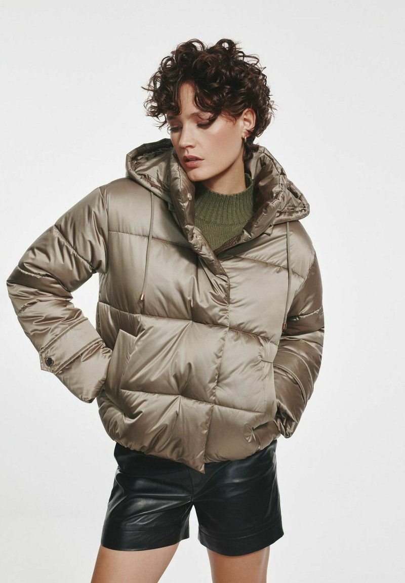 Zielona kurtka puchowa w kolorze oliwkowym o błyszczącej, pikowanej fakturze. Posiada zamek błyskawiczny z przodu, dużą kieszeń oraz boczne kieszenie. Połączona z czarnymi krótkimi spodniami.