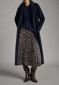 Marineblauwe lange jas met een scherpe kraag, geribde marineblauwe trui en geplooide bruin met luipaardprint rok. Kniehoge bruine hoge hakken maken de look compleet.