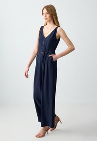 Jumpsuit senza maniche blu navy con scollo a V profondo, vita cinchata e tasche laterali. Presenta passanti per cintura e un tessuto leggero per il comfort.