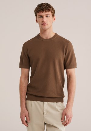 Jeune homme aux cheveux bouclés portant une chemise marron texturée à manches courtes et un pantalon beige clair, debout devant un fond uni.