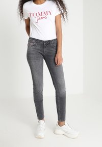 Camiseta blanca con el logotipo rojo "TOMMY Jeans", combinada con jeans ajustados grises y zapatillas blancas, de pie contra un fondo neutro.