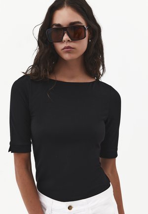 Vrouw met lang donker haar, die grote vierkante zonnebril draagt, een zwart nauwsluitend topje met halflange mouwen en een witte broek tegen een effen achtergrond.