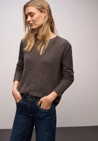 Grauer Strickpullover mit langen Ärmeln und rundem Ausschnitt, kombiniert mit dunkelblauen Jeans. Der Stoff wirkt strukturiert und weich.