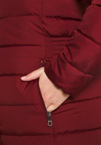 Bordeaux puffjakke med kviltad textur och en dragkedjeficka. Materialet verkar slätt, med elastiska sidopaneler för bättre passform.