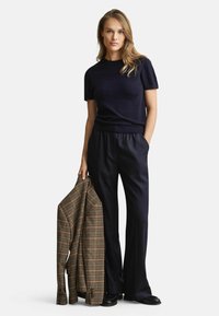 Marineblauwe gebreide top met korte mouwen gecombineerd met donkere, wijde broek, terwijl een bruine geplaid blazer wordt vastgehouden, wat texturen en contrasterende kleuren benadrukt.