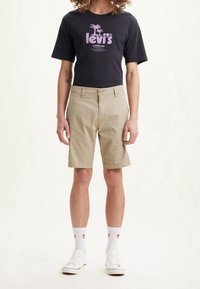 Schwarzes T-Shirt mit pinkem Logo, khakifarbene Shorts, weiße Sneakers und weiße Socken mit roten Akzenten. Lässiges, kurzärmeliges Outfit.
