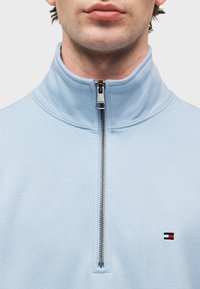 Ανοιχτό μπλε φούτερ με φερμουάρ, διαθέτει ψηλό γιακά και ασημένιο φερμουάρ. Μικρό λογότυπο κεντημένο στο στήθος σε κόκκινο, άσπρο και ναυτικό μπλε.