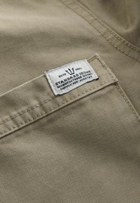 Khaki fabric with a textured surface, featuring a white label sewn on a pocket—text includes "ESTD 1982," "STANDARD ISSUE," and supply reference.