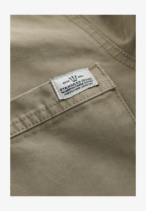 Khakifarbenes Gewebe mit strukturierter Oberfläche, versehen mit einem weißen Etikett, das auf einer Tasche genäht ist – der Text umfasst "ESTD 1982", "STANDARD ISSUE" und eine Lieferreferenz.