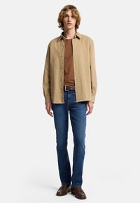 Beżowa koszula z długim rękawem na guziki nałożona na brązowy T-shirt, zestawiona z niebieskimi dżinsami i brązowymi mokasynami. Model stoi na jednolitym tle.