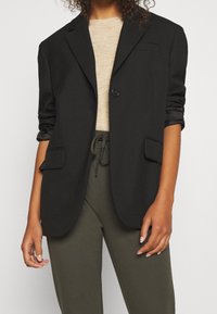 Donna che indossa un blazer nero sopra un maglione beige e pantaloni verde oliva con coulisse, con le maniche del blazer risollevate.