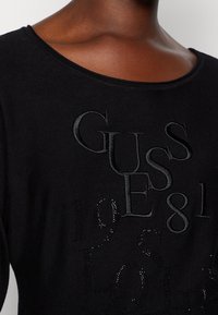 Sudadera negra con letras en relieve de "GUESS" y una textura suave. Los detalles notables incluyen adornos de cristal integrados en el diseño.