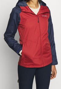 Röd och marinblå lätt jacka med dragkedja fram, huva och kontrasterande paneler. Har en slät yta och Columbia-logotypen på bröstet.
