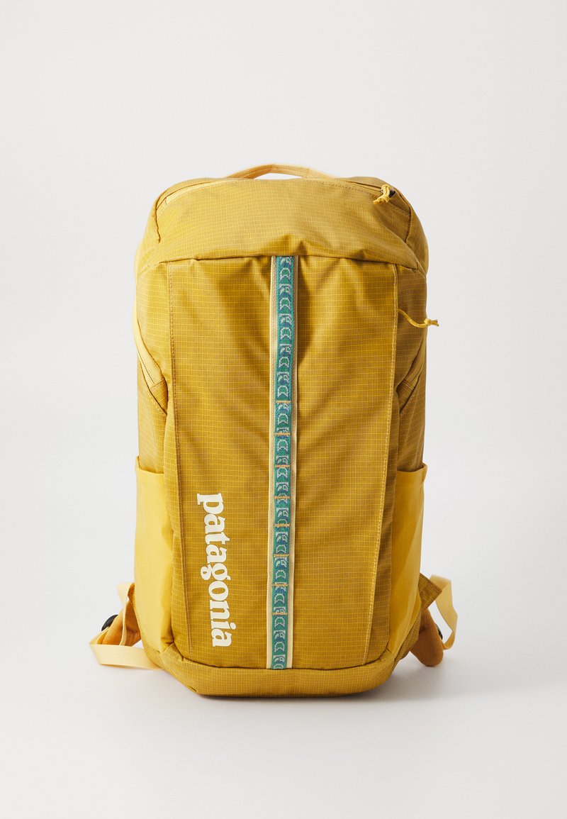Mochila amarela com um padrão em grelha, apresentando uma faixa vertical verde e um logotipo branco. Inclui bolsos laterais e alças ajustáveis.