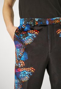 Sorte bukser med en livlig sommerfugleprint i blå, orange og rød, med en flad front og et bælte med et matchende mønster.