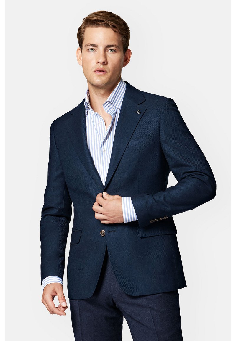 Granatowa marynarka o dopasowanym kroju, zapięcie na dwa guziki, klapki z wcięciem oraz delikatna tekstura. W zestawie z niebieską koszulą w białe paski pod spodem.