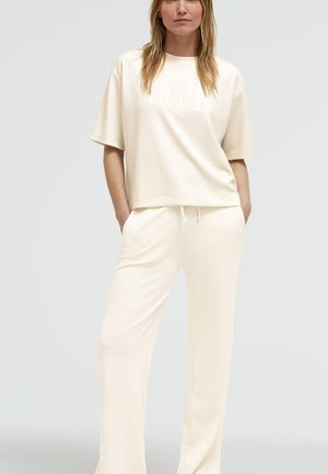 Frau trägt locker sitzendes, cremefarbenes Oberteil mit kurzen Ärmeln und passende weite Hose, steht mit Händen in den Taschen vor neutralem Hintergrund.